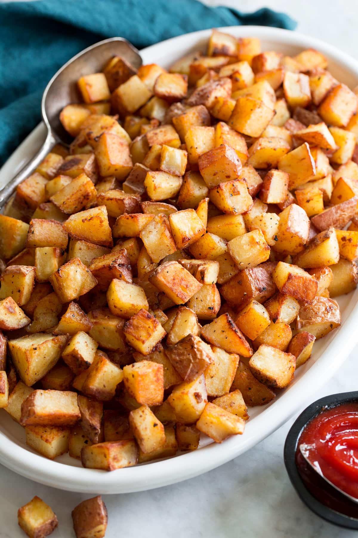 Breakfast Potatoes Photo: Breakfast Potatoes on a white oval serving plater with a blue cloth and silver spoon resting to the side. Sriracha ketchup is shown as a serving suggestion.
