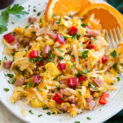 Photo: Serving of breakfast hash on a white ceramic plate with oranges to the side. Hash is made with shredded hash browns, ham, eggs, bell pepper, parsley and onion.