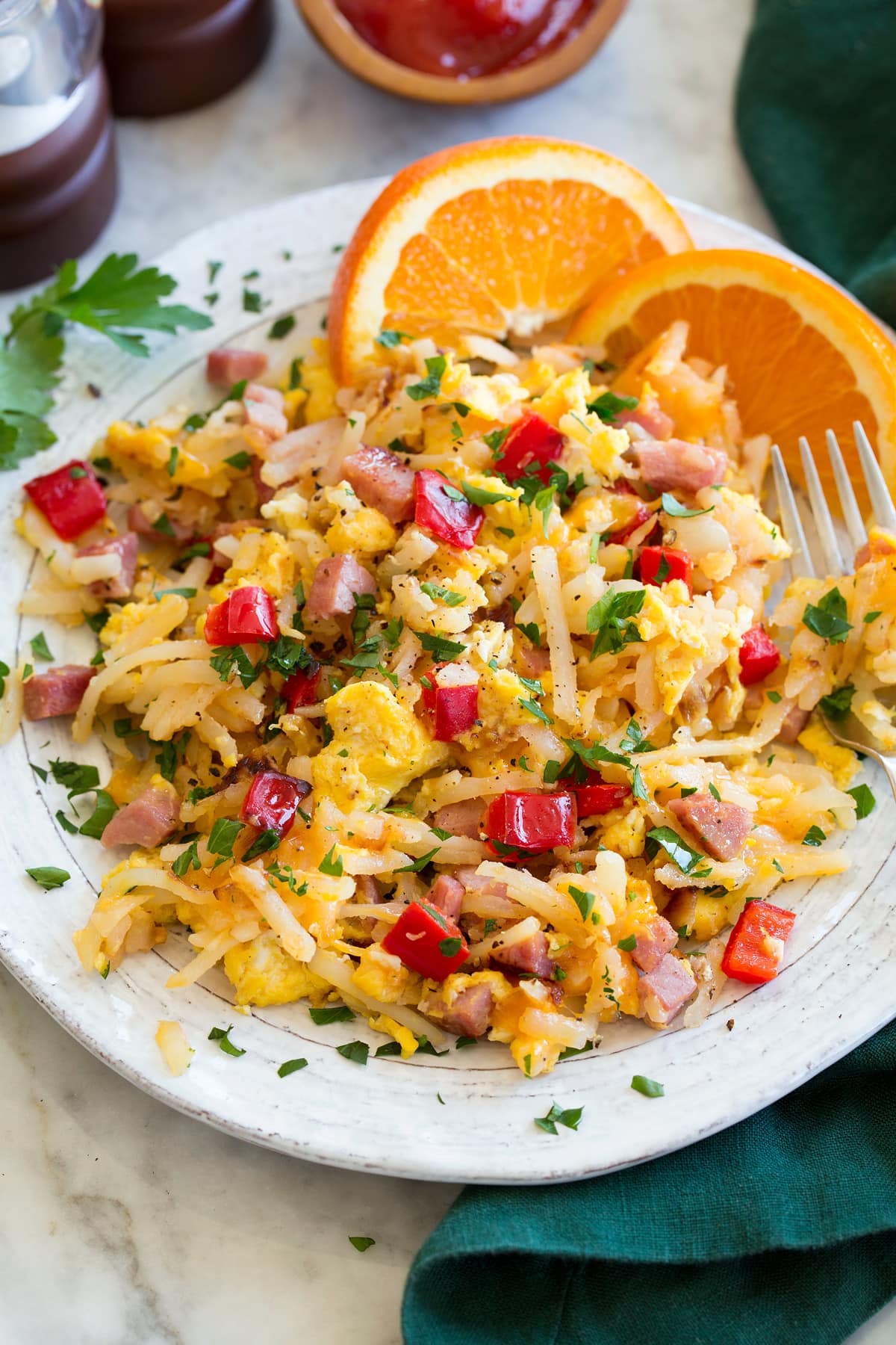 Photo: Serving of breakfast hash on a white ceramic plate with oranges to the side. Hash is made with shredded hash browns, ham, eggs, bell pepper, parsley and onion.