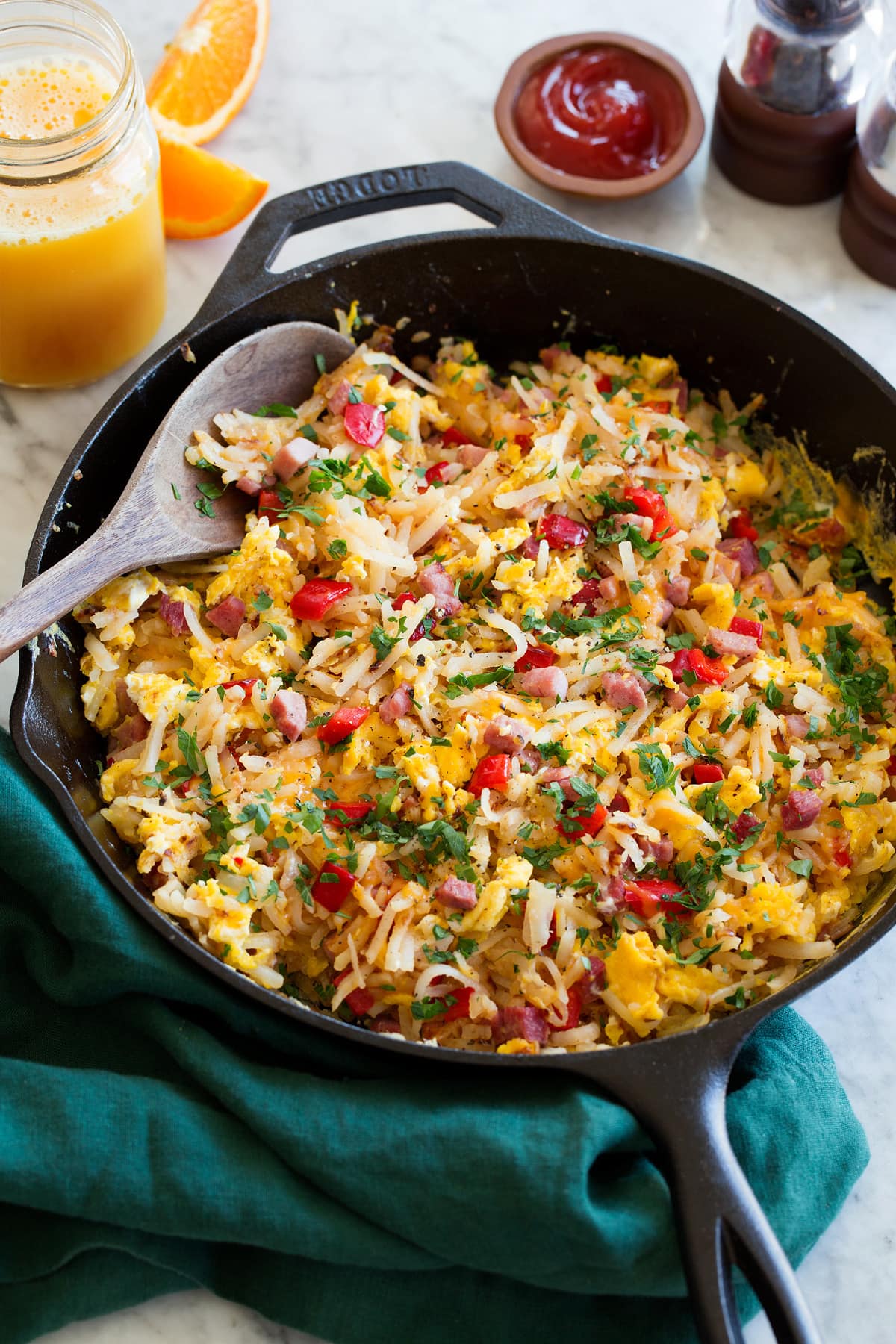 Photo: Egg and hash brown breakfast hash shown in a black cast iron skillet resting on a marble surface. A green cloth is shown to the side. Orange juice and ketchup are shown as serving suggestions.