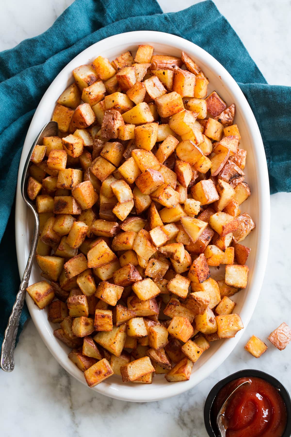 Breakfast Potatoes Photo: Diced breakfast potatoes shown from above on a white oval serving platter with a marble surface below and a blue cloth.