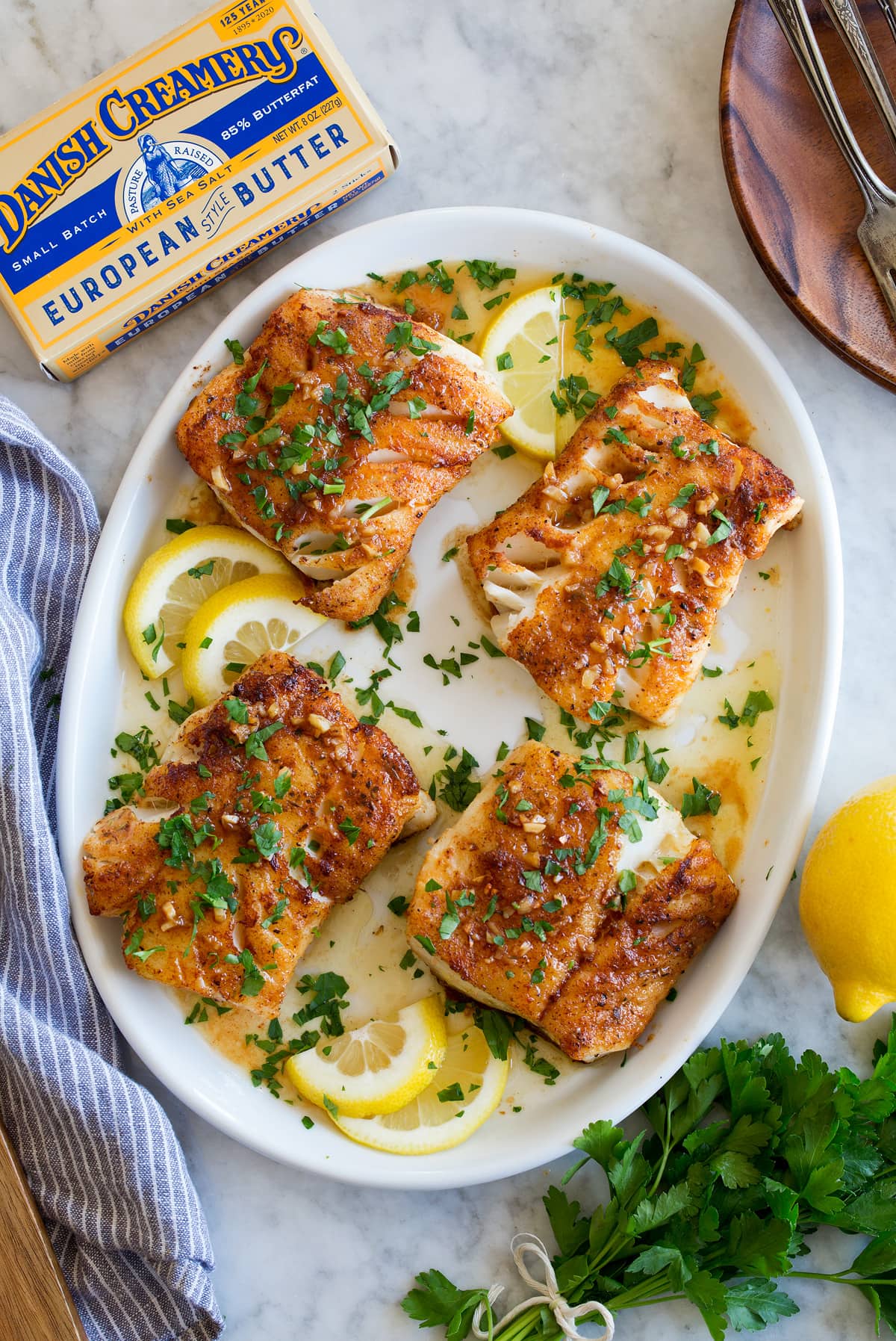 Four cod fillets shown overhead on a white oval platter. They are covered in a sauce and garnished with parsley. A package of butter and fresh parsley are shown to the side.