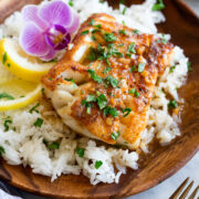 Four cod fillets shown overhead on a white oval platter. They are covered in a sauce and garnished with parsley. A package of butter and fresh parsley are shown to the side.