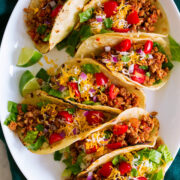 Photo: 7 ground turkey tacos in a row shown on a white oval platter from above. Tacos are filled with meat, tomatoes, lettuce, cheese and red onion.