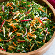 Photo: Kale salad with sliced apples, carrots, red onions, white cheddar and almonds shown in a wooden bowl over a marble surface. A red cloth is shown to the side and wooden salad spoons.