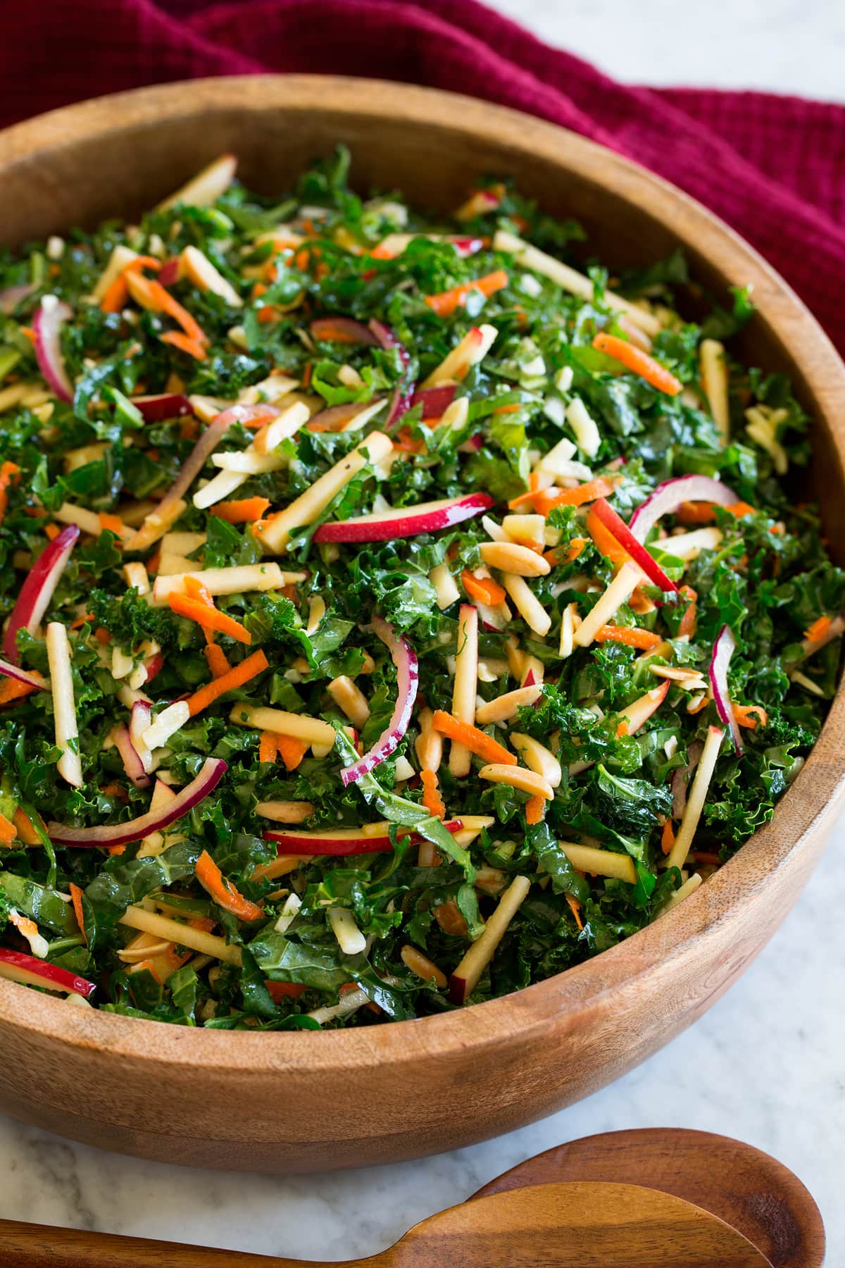 Photo: Kale salad with sliced apples, carrots, red onions, white cheddar and almonds shown in a wooden bowl over a marble surface. A red cloth is shown to the side and wooden salad spoons.