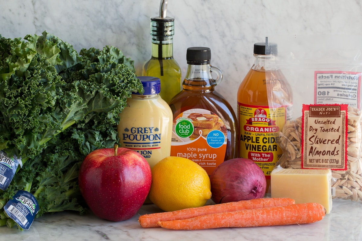 Photo showing ingredients used to make kale salad. Includes kale, apples, lemon, red onion, carrots, white cheddar, maple syrup, apple cider vinegar, dijon mustard, olive oil and slivered almonds.
