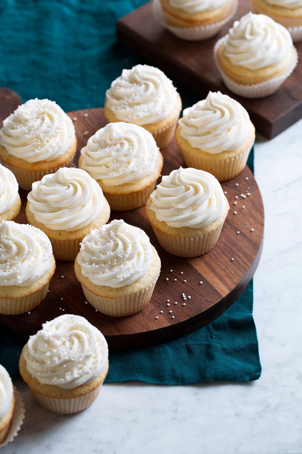 A batch of homemade vanilla cupcakes shown on a wooden serving platter over a blue cloth.