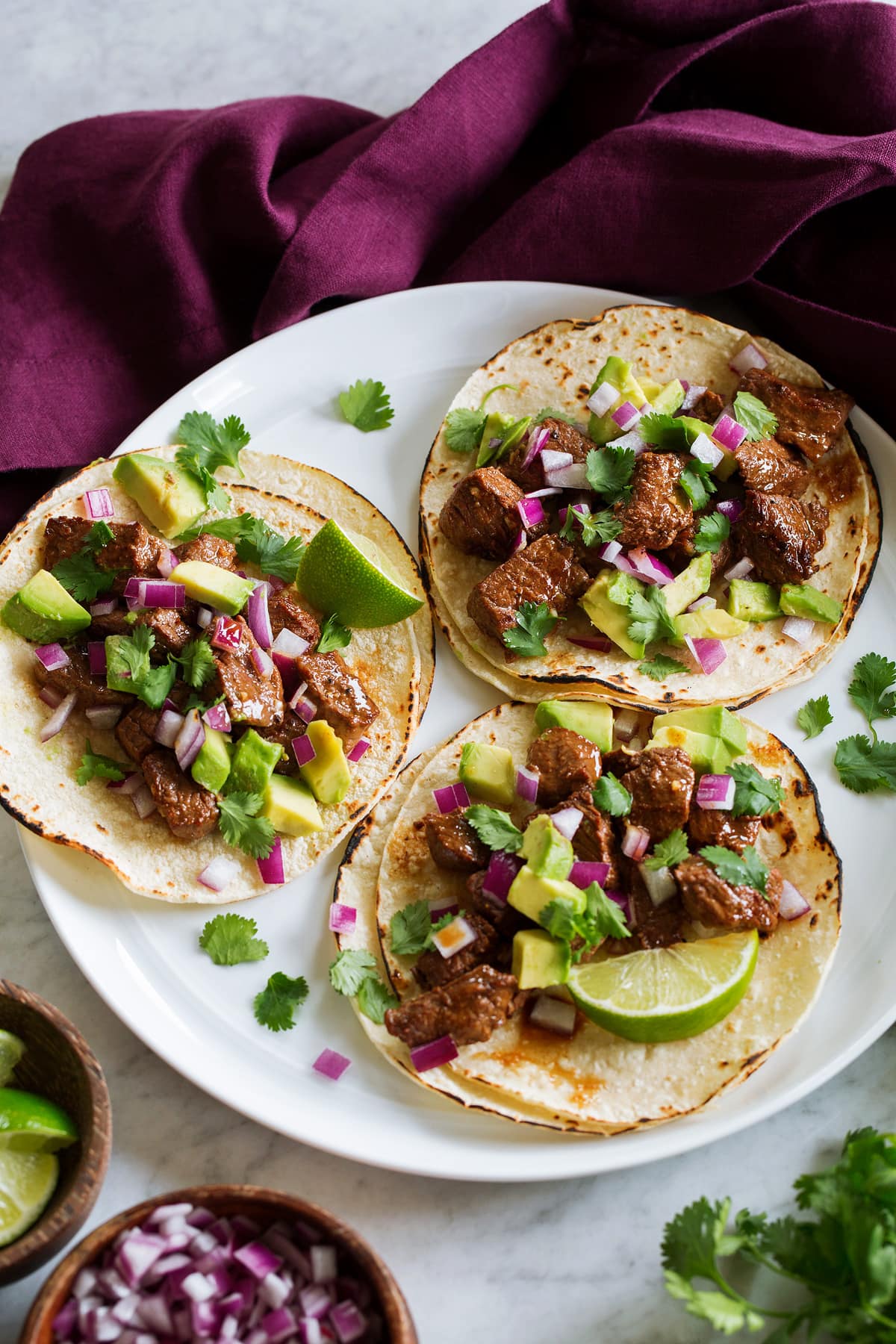Steak Tacos Three steak tacos on a white serving plate with a purple cloth to the side, shown from a side angle.