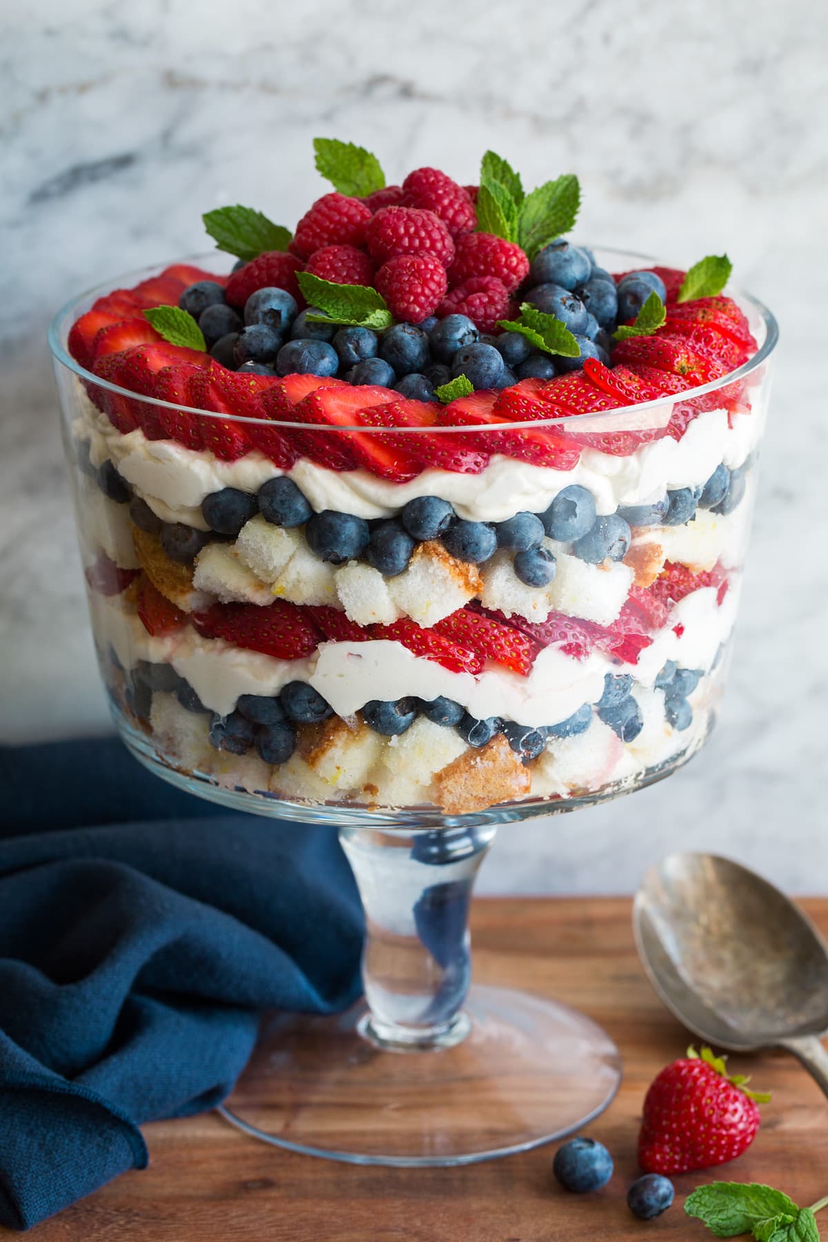 Layered trifle in a glass trifle dish. It includes cubes of angel food cake, blueberries, strawberries, raspberries and a cream cheese whipped cream topping.