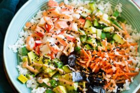 California roll sushi bowl shown in a blue bowl from a side angle. Bowl is resting on a wooden plate over a marble surface.