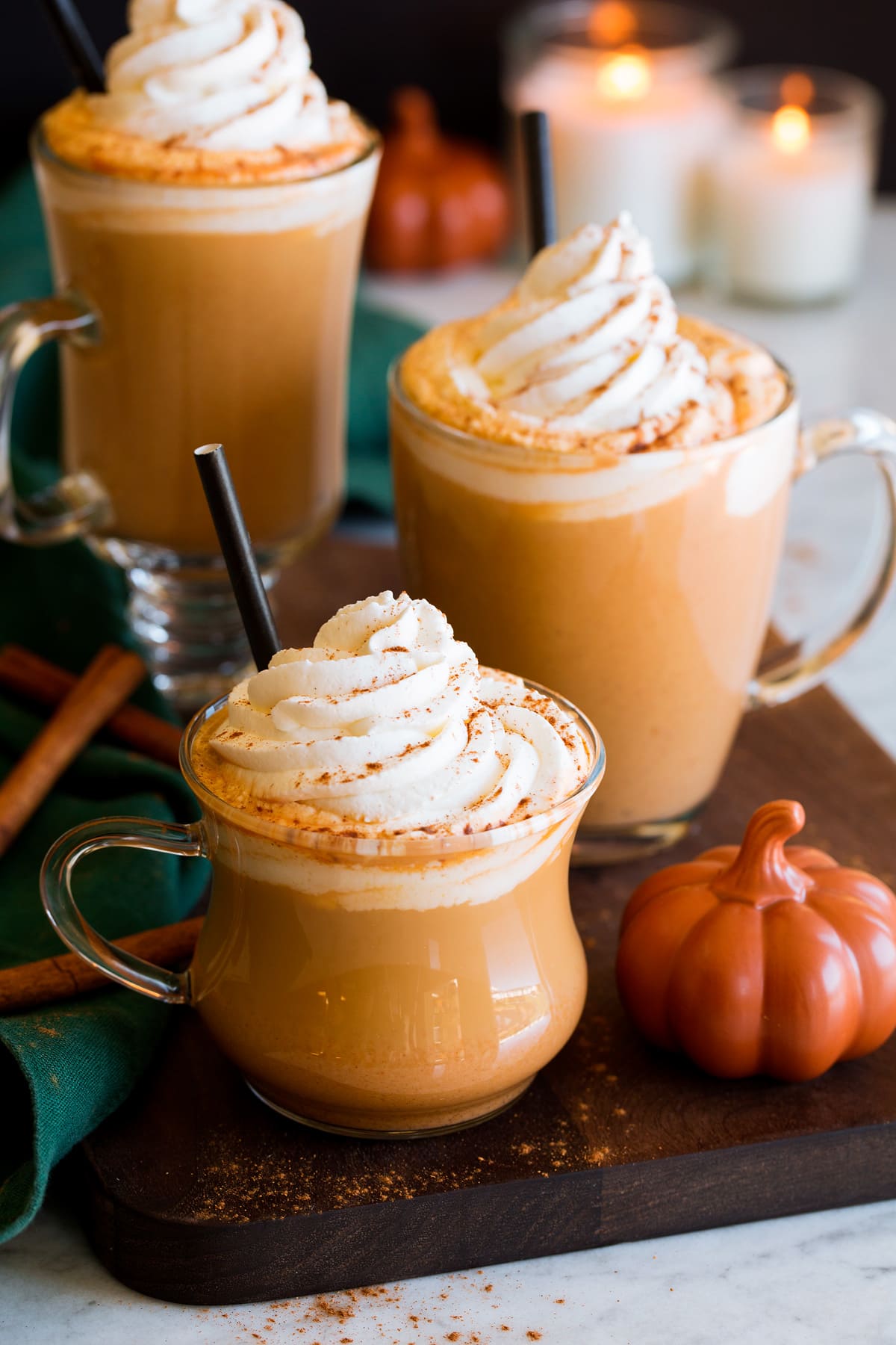 Pumpkin Spice Latte Three pumkpin spice lattes with a decorative pumpkin to the side, cinnamon sticks and candles in the background. Lattes are topped with whipped cream and pumpkin spice.