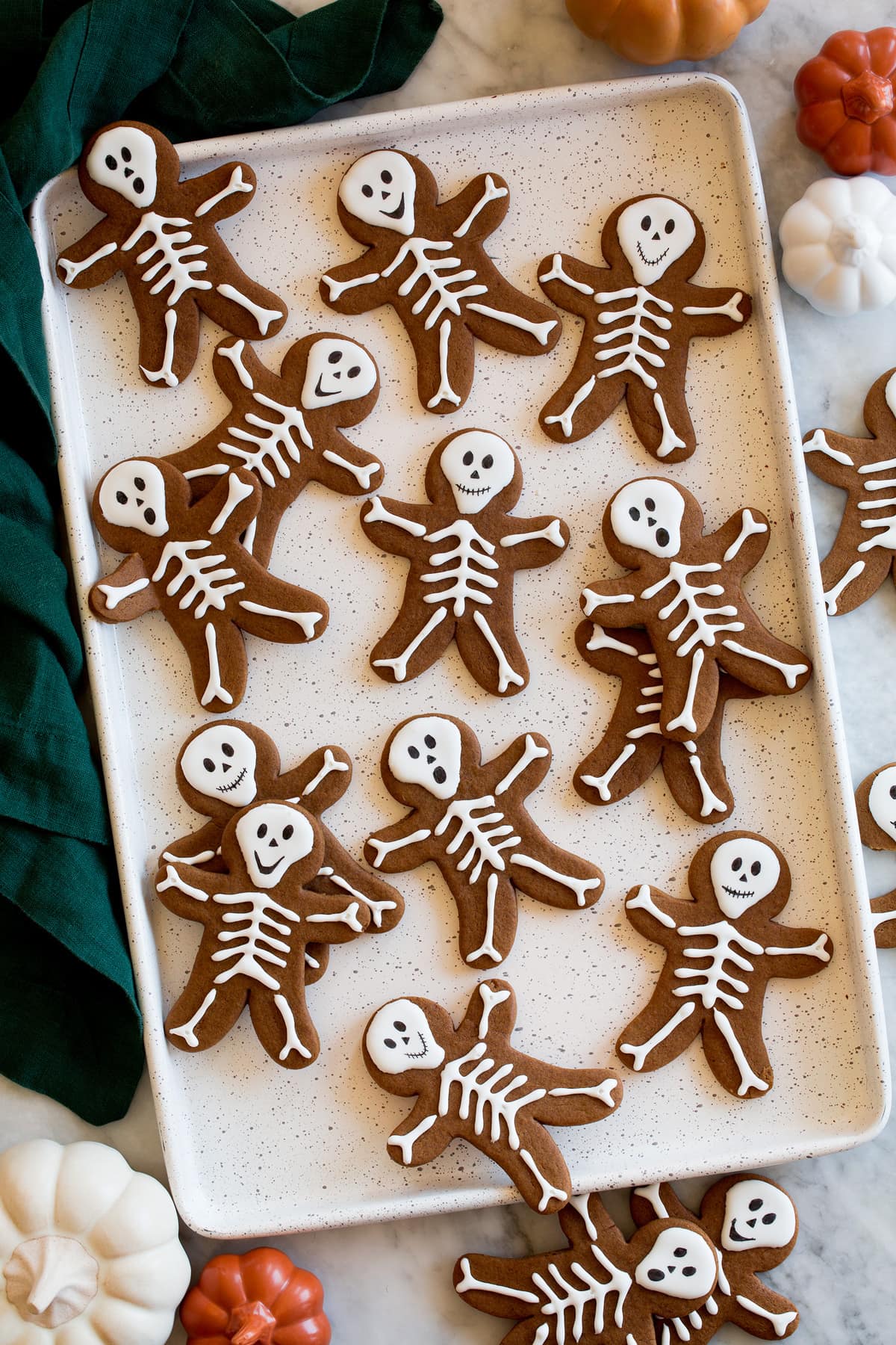 Skeleton Gingerbread Cookies Skeleton gingerbread cookies on a white baking sheet with a green cloth and decorative pumpkins to the side.