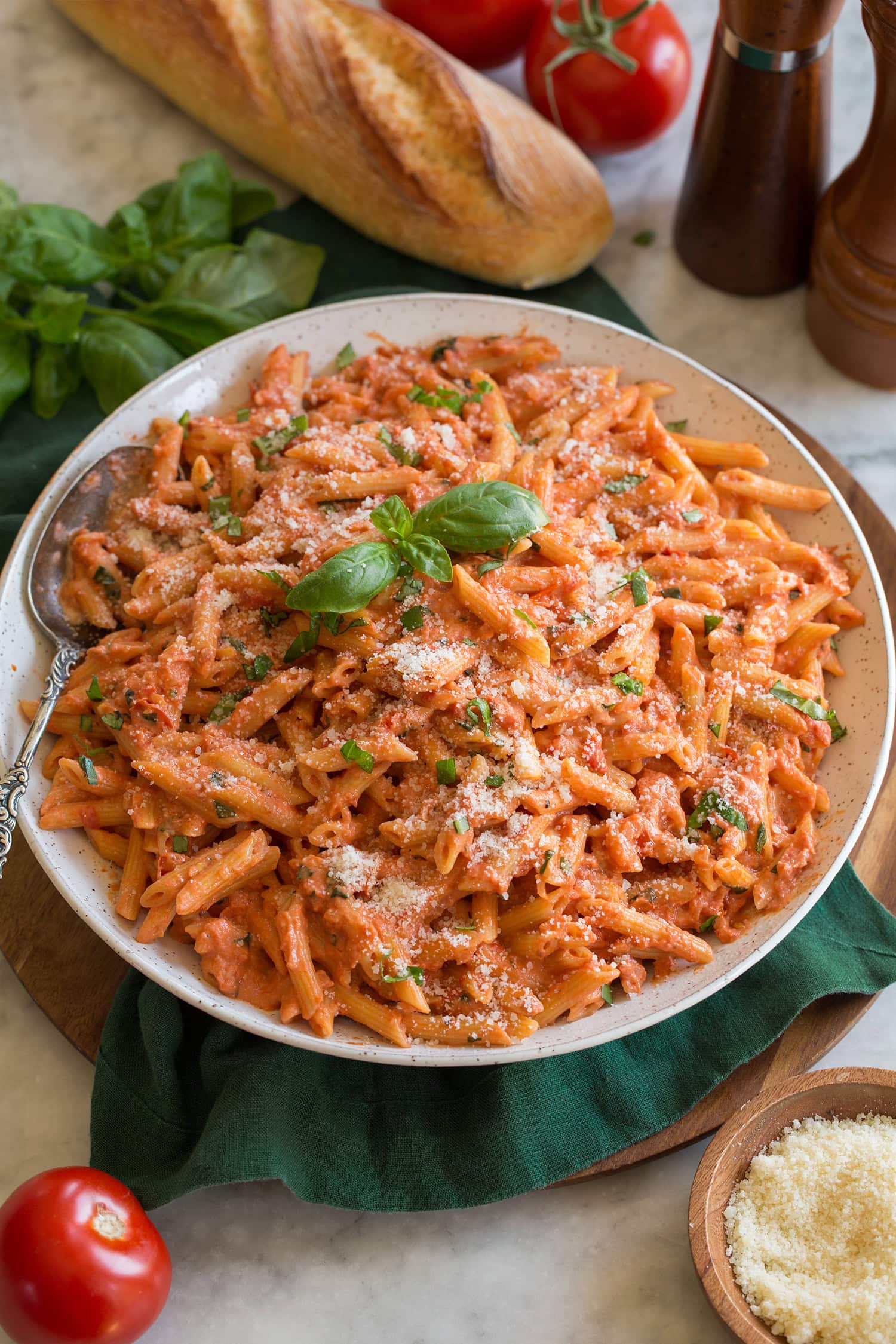 Creamy tomato pasta garnished with parmesan. Shown served in a large white bowl over a green cloth on a marble surface. A baguette and whole tomatoes are shown to the side.