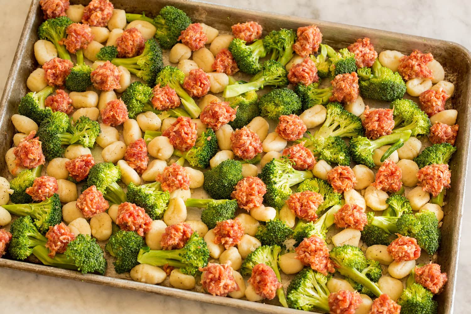 Raw sausage crumbles layered over broccoli and gnocchi tossed together on a sheet pan.