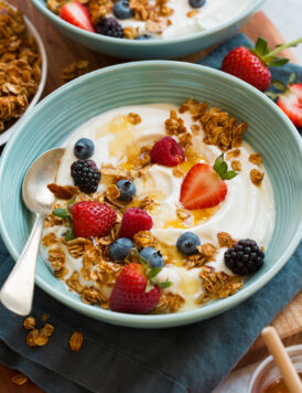 Homemade yogurt with fresh berries, granola and honey in a teal bowl over a blue cloth.