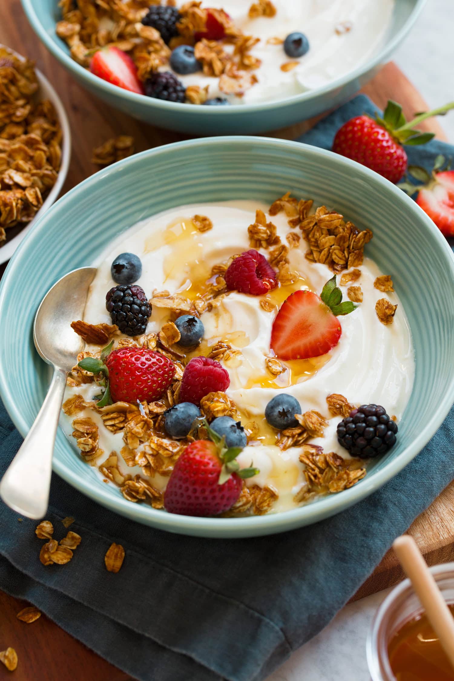 Homemade yogurt with fresh berries, granola and honey in a teal bowl over a blue cloth.