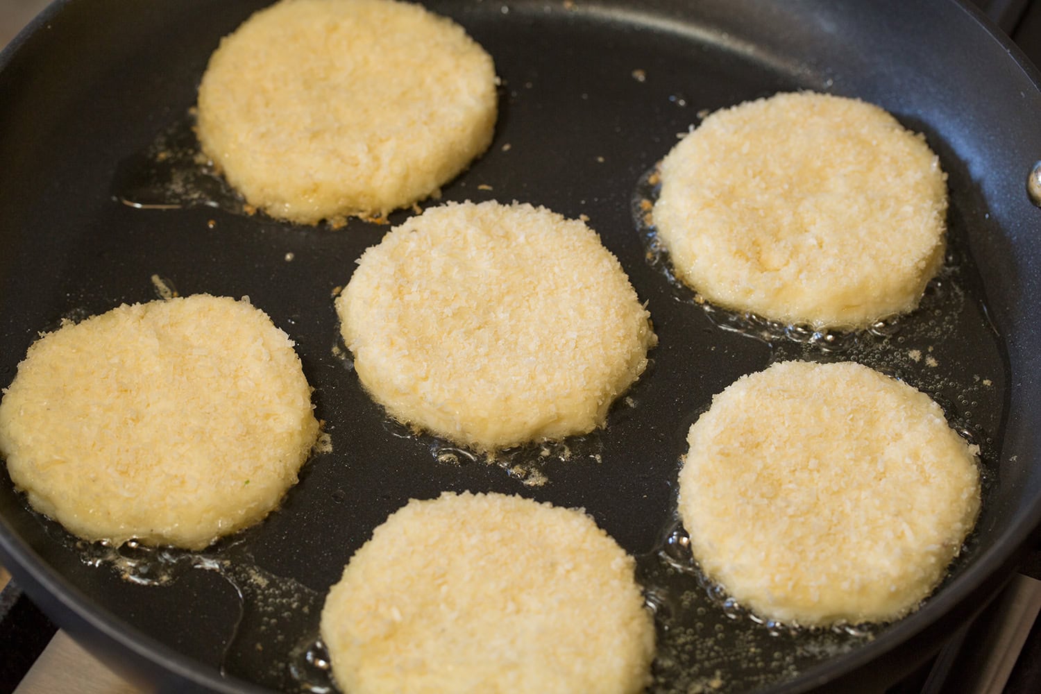 Mashed Potato Cakes Potato cakes being fried in oil in a dark non-stick skillet.