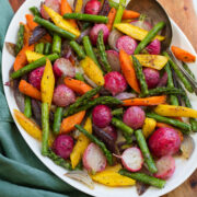 Colorful roasted spring vegetables on a white oval platter on a wooden board with a teal cloth to the side.