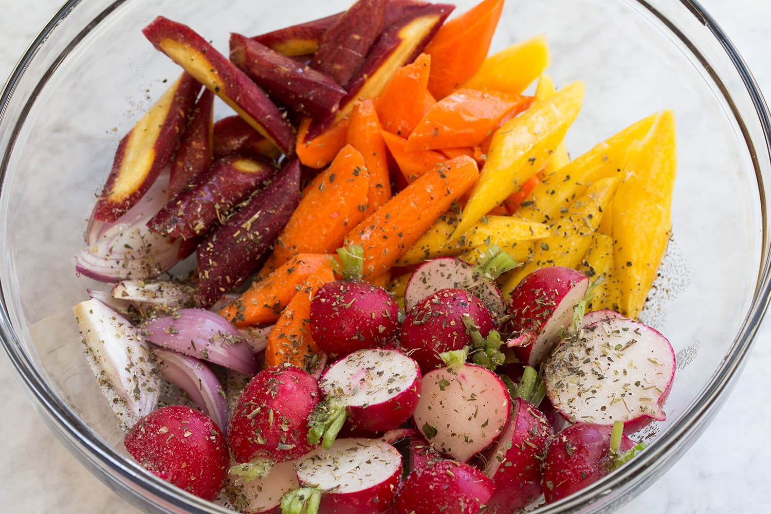 Raw chopped vegetables in a glass bowl with seasonings and oil before tossing.