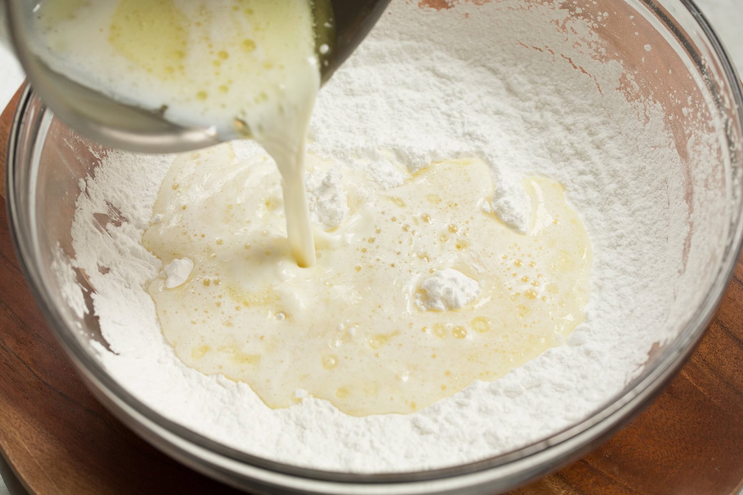 Brazilian Cheese Bread (Pão de Queijo) Scalded oil and milk mixture being poured into tapioca flour in a glass mixing bowl.