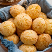 Homemade Brazilian cheese bread in a wooden bowl with a blue cloth underneath the bread. Grated cheese is in the background.
