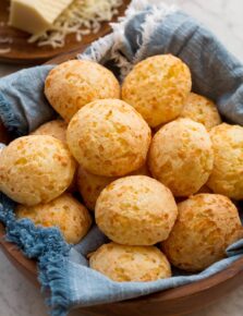 Homemade Brazilian cheese bread in a wooden bowl with a blue cloth underneath the bread. Grated cheese is in the background.