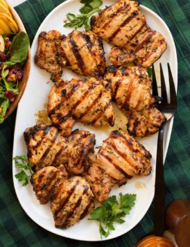 Marinated grilled chicken thighs shown on a white oval platter from above. Platter is resting on a green and blue checked cloth and a pear cranberry spinach salad is shown to the side.