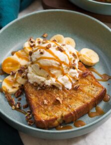 Close up photo of banana bread sundae. Shows skillet browned banana bread slice with vanilla ice cream scoop on top, banana slices in the back. And everything is drizzled with caramel sauce.