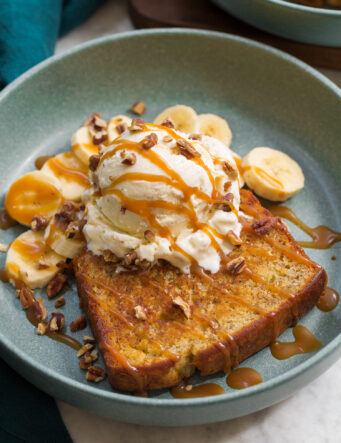 Close up photo of banana bread sundae. Shows skillet browned banana bread slice with vanilla ice cream scoop on top, banana slices in the back. And everything is drizzled with caramel sauce.