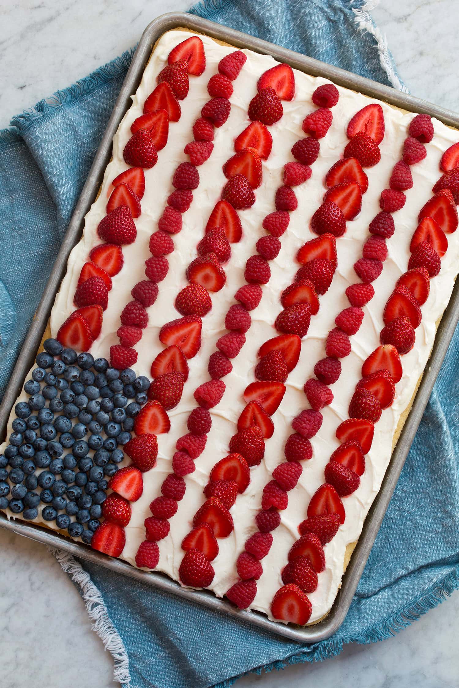 American flag cake made in a sheet pan. Shown topped with frosting, sliced strawberries, blueberries and raspberries.