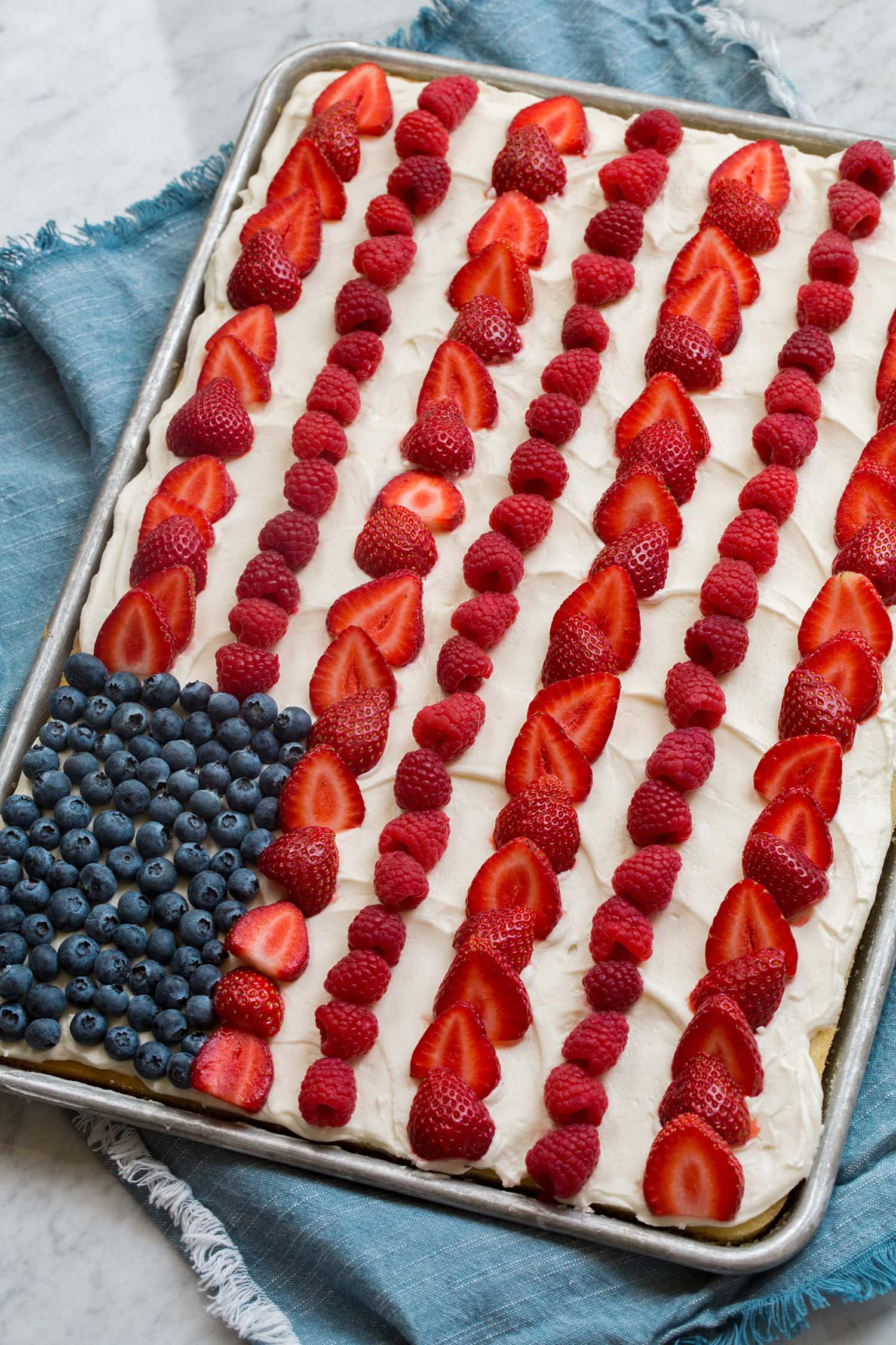American flag cake with fresh berries and cream cheese frosting shown in a baking sheet on a blue cloth over a marble surface.