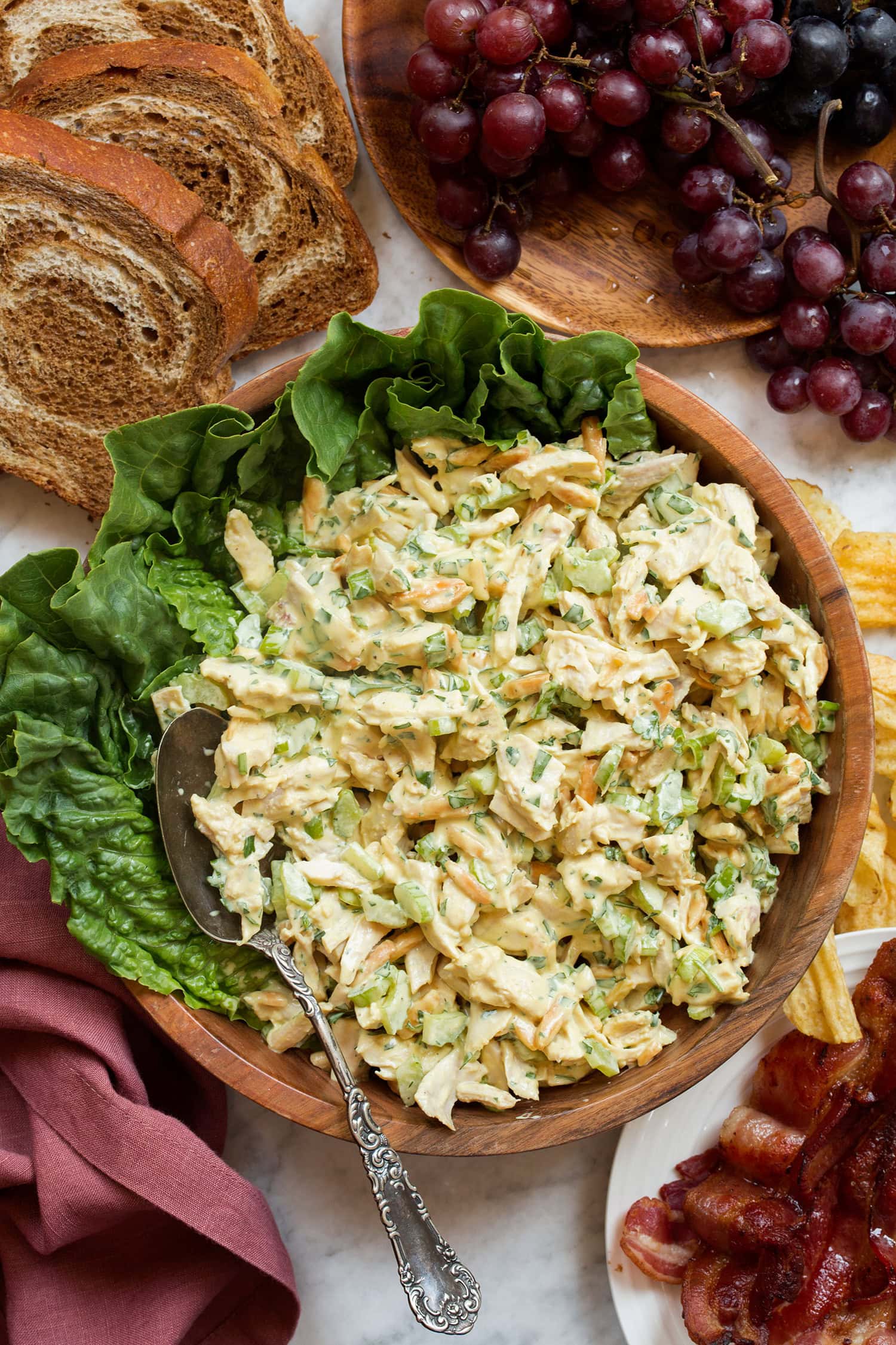 Bowl of of chicken salad with honey mustard sauce, shown in a wooden bowl with lettuce leaves. Grapes, bacon and bread slices are laid decoratively around the bowl.