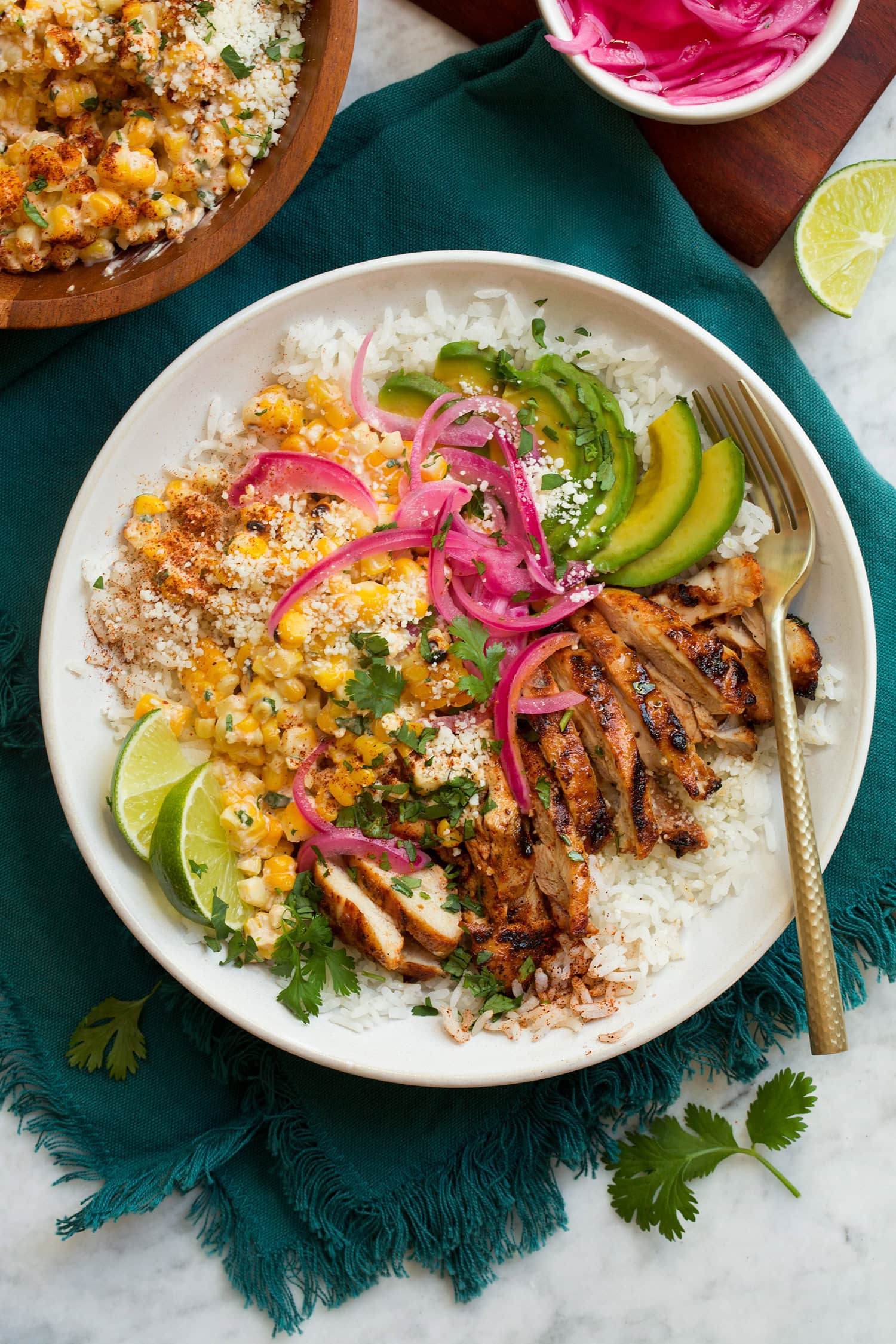 Overhead image of street corn and chicken rice bowl on a blue cloth on a marble surface.
