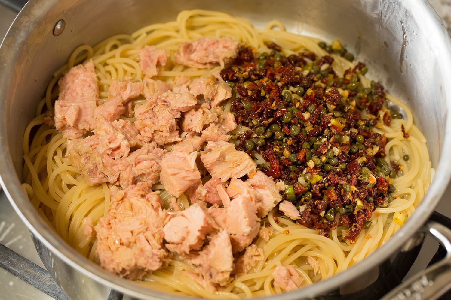 Tuna and sun dried tomato caper mixture added to cooked spaghetti in a stainless steel pot.