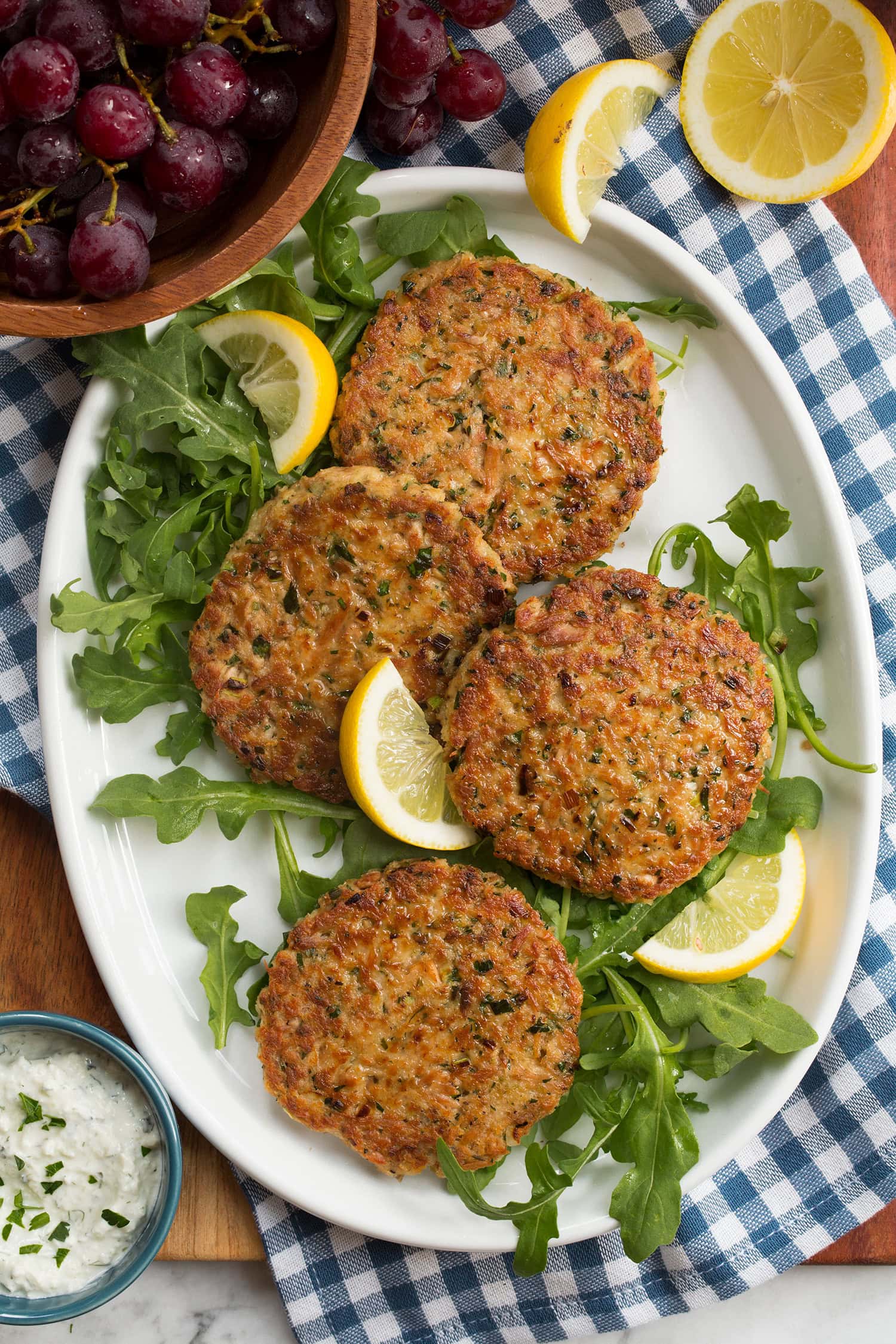 Four tuna patties shown from overhead on a white oval serving platter over a blue checkered cloth on a wooden platter.