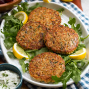 Tuna Patties shown on a platter over a blue checkered cloth. Lemons and arugula are shown around the tuna patties.