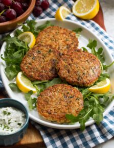 Tuna Patties shown on a platter over a blue checkered cloth. Lemons and arugula are shown around the tuna patties.