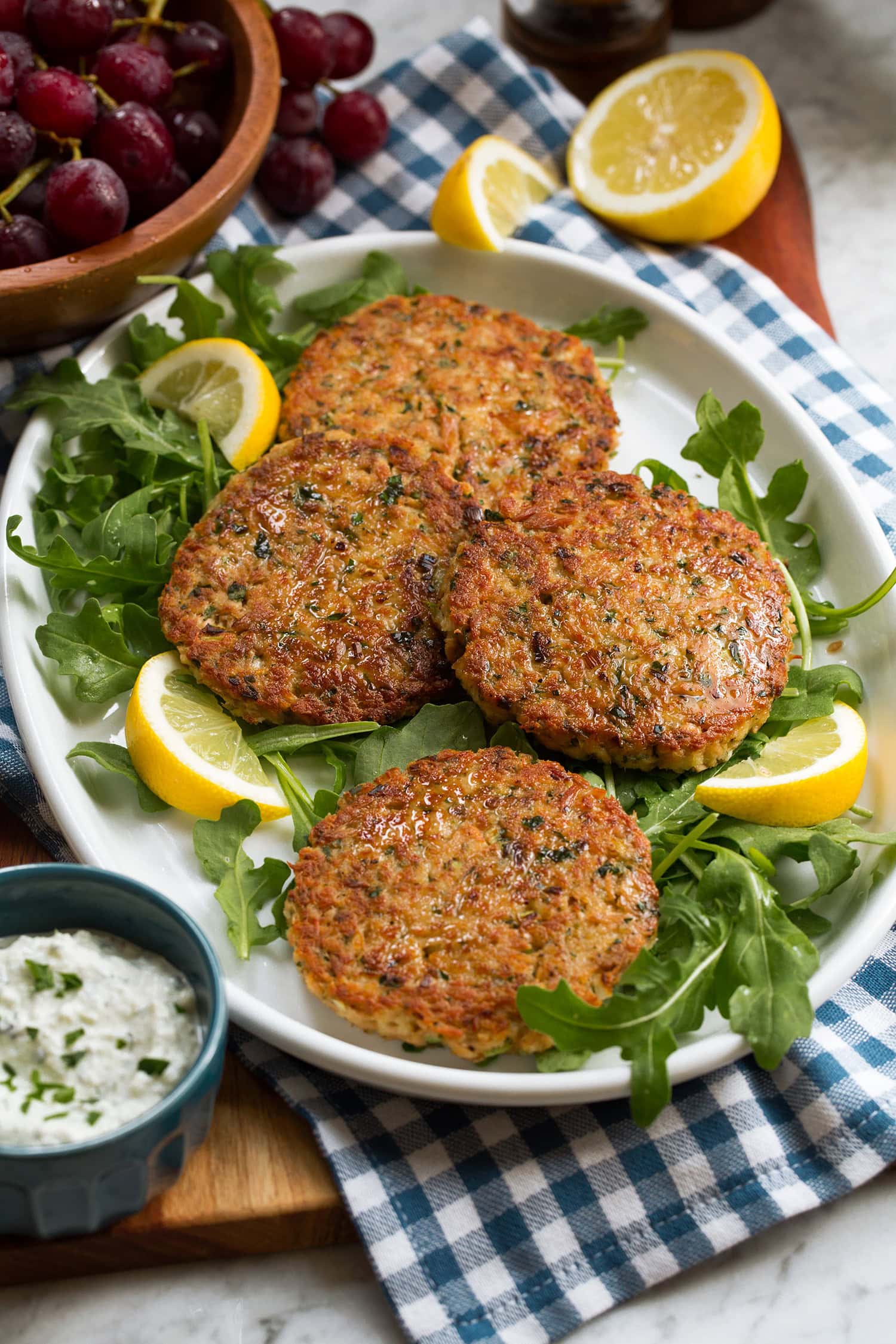 Tuna Patties shown on a platter over a blue checkered cloth. Lemons and arugula are shown around the tuna patties.