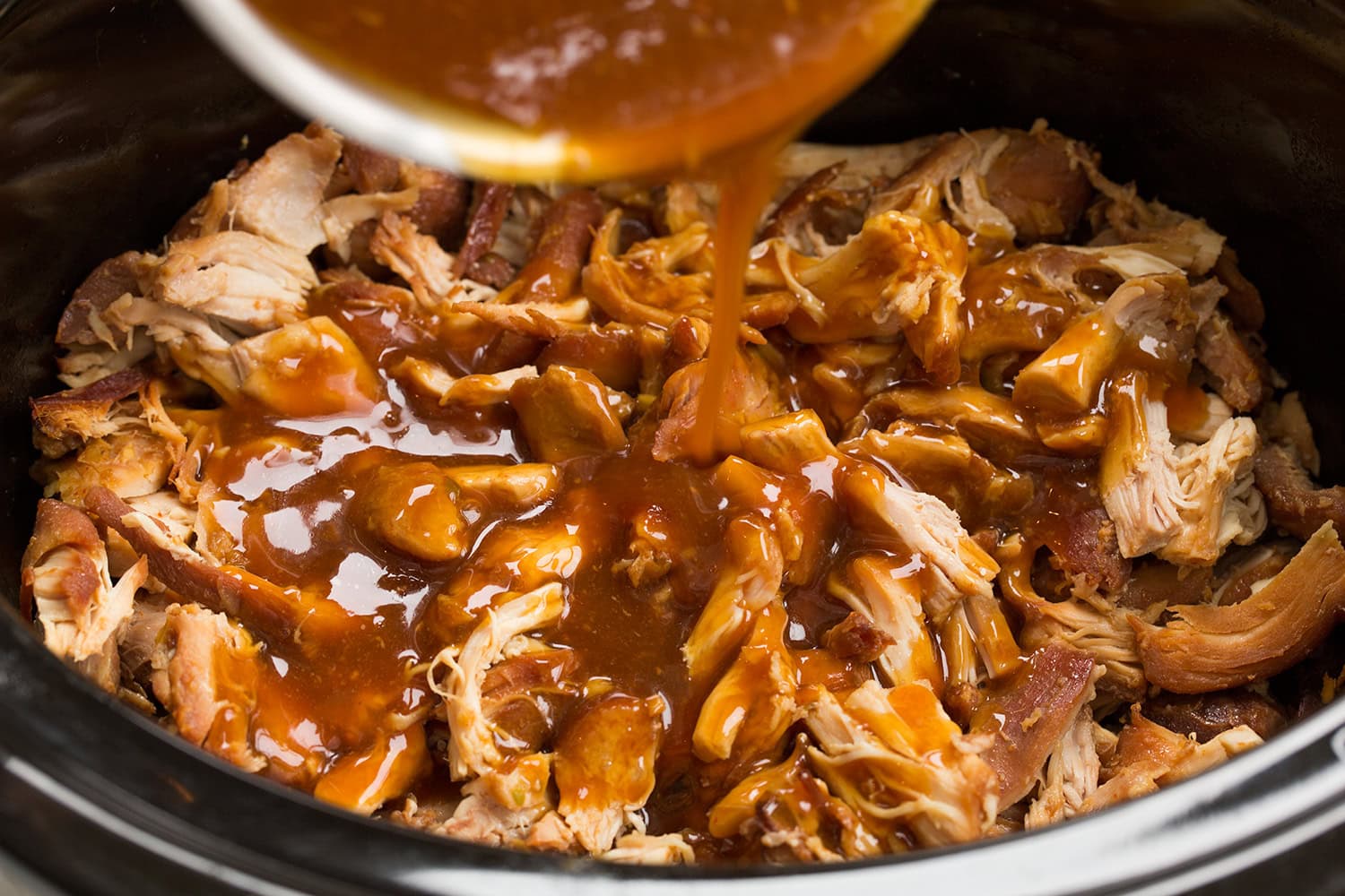 Honey garlic sauce being poured over shredded chicken in a slow cooker.